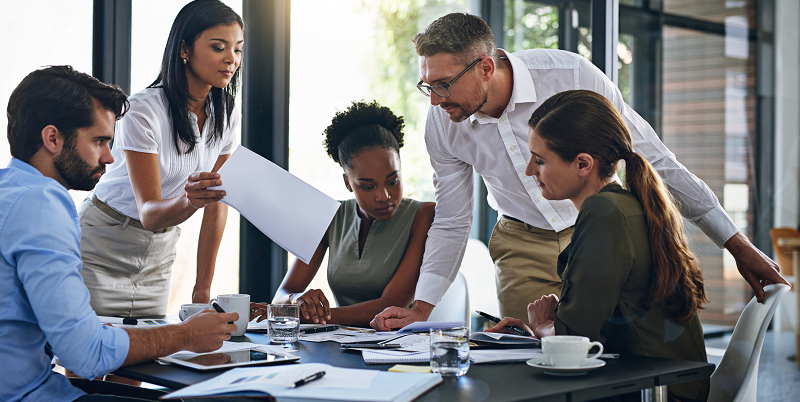 A diverse group of employees stand around a table reviewing documents.