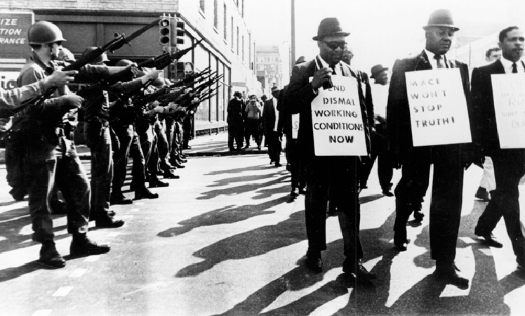 Black sanitation workers participate in a strike in Memphis in 1968 as National Guard members look on. One worker carries a sign that reads “End dismal working conditions now.”