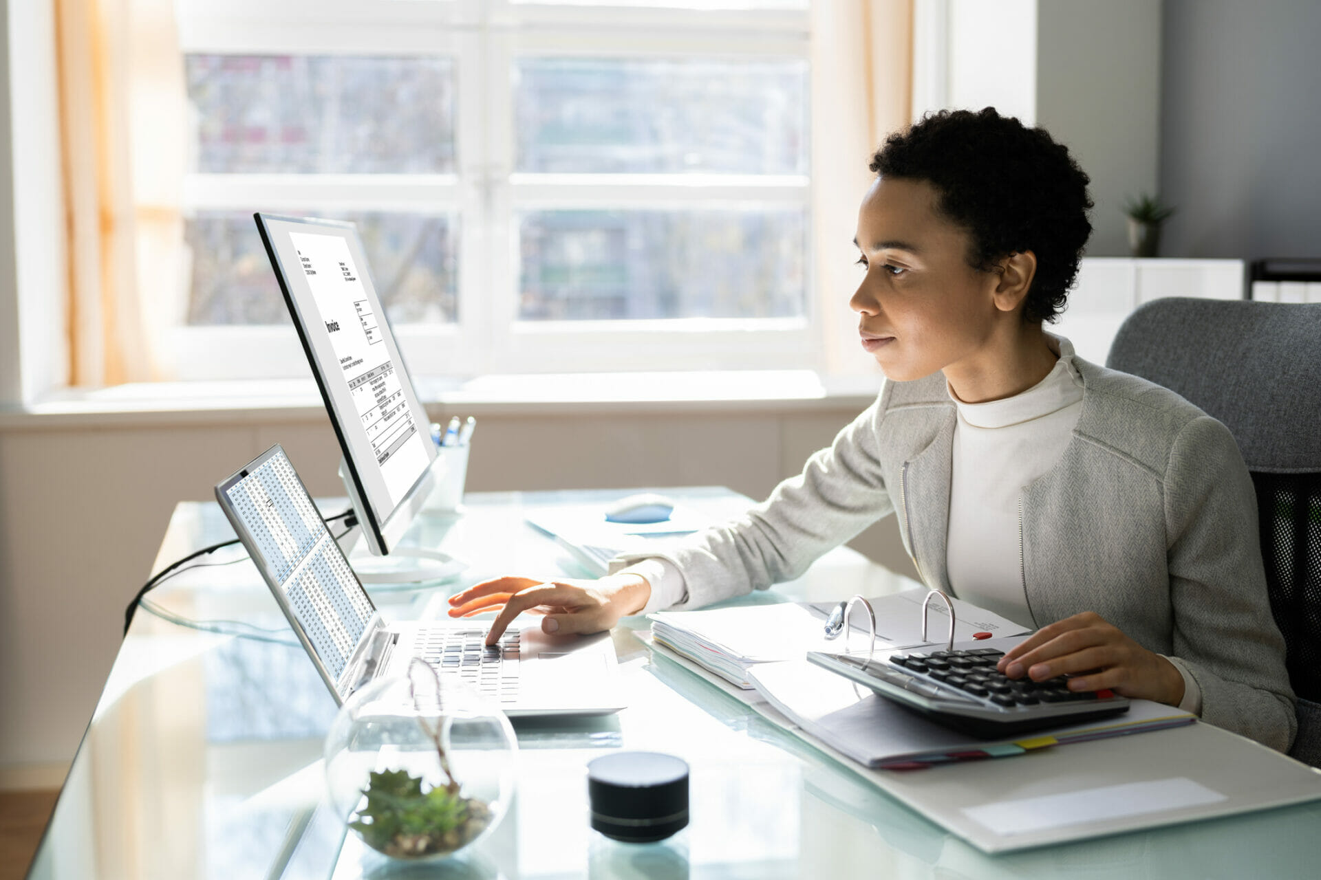 Professionally dressed woman seated at a desk with computers and notebooks.