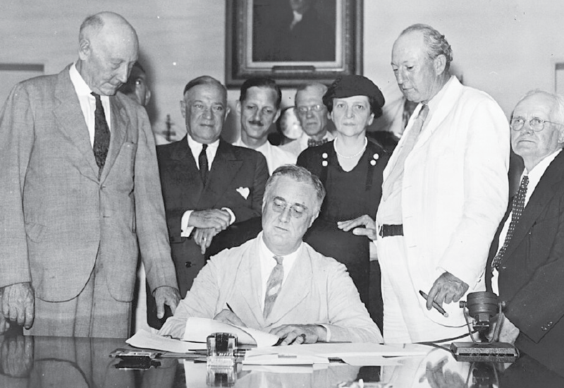 Black and white photo: President Franklin Roosevelt signs the Social Security Act, flanked by supporters, including Secretary Frances Perkins.