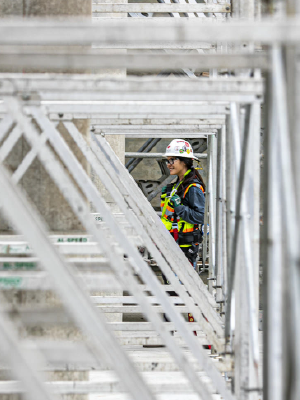 A woman in a hardhat and protective gear stands on scaffolding, overlooking a construction site.