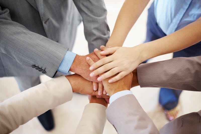 Group of colleagues dressed in various business casual attire joining hands in the middle to show unity.