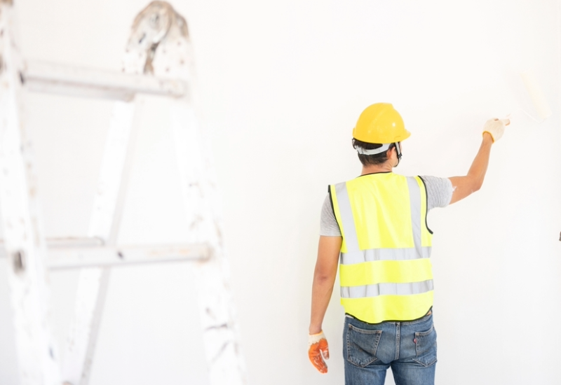 A worker wearing a high visibility vest and hardhat, seen from behind, touches up a white wall with a paint brush.