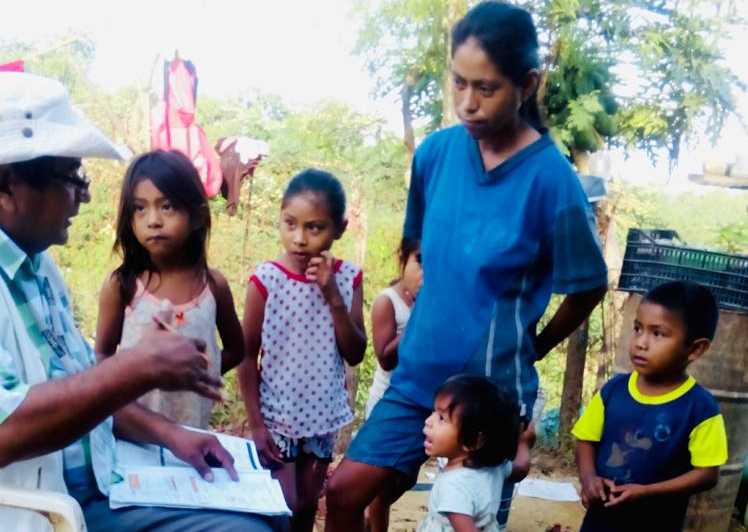 A man holding paperwork and a pencil speaks outdoors with a woman who is surrounded by several small children.