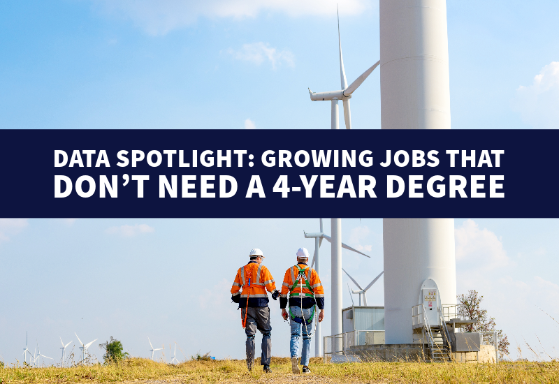 Two workers wearing fall protection gear walk toward large wind turbines in a field.