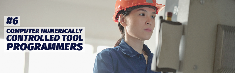 Woman wearing a hardhat stands at a computer-type device in an industrial environment.