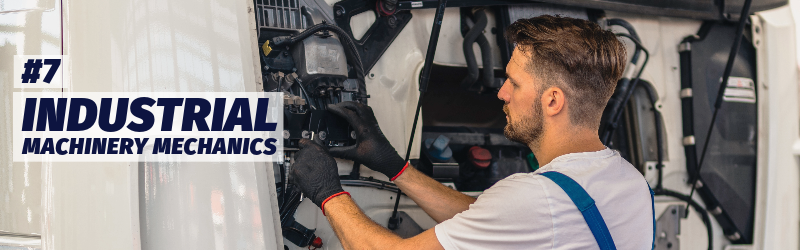 A man wearing overalls and work gloves looks at a panel under the hood of a large truck.