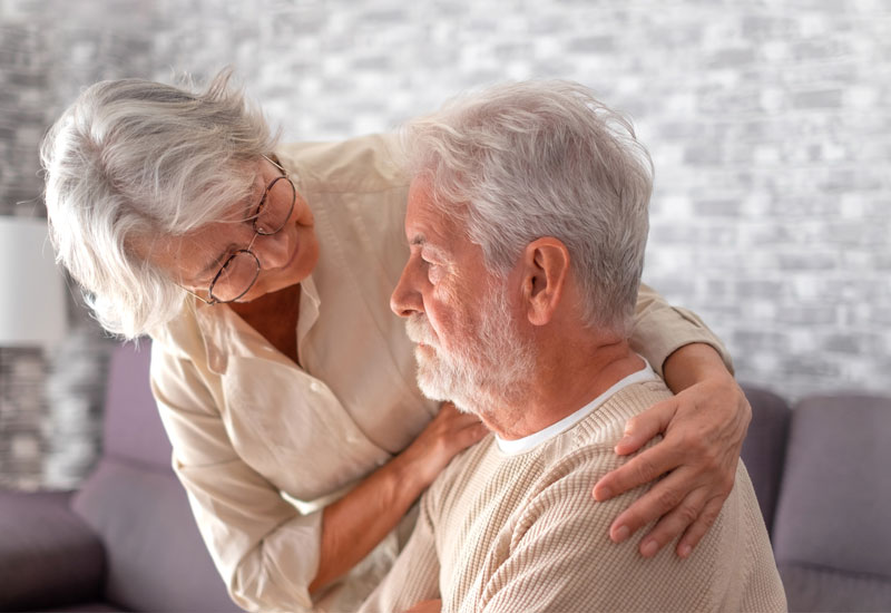 An older woman leans toward an older man who is seated. Her arms enfold him supportively.