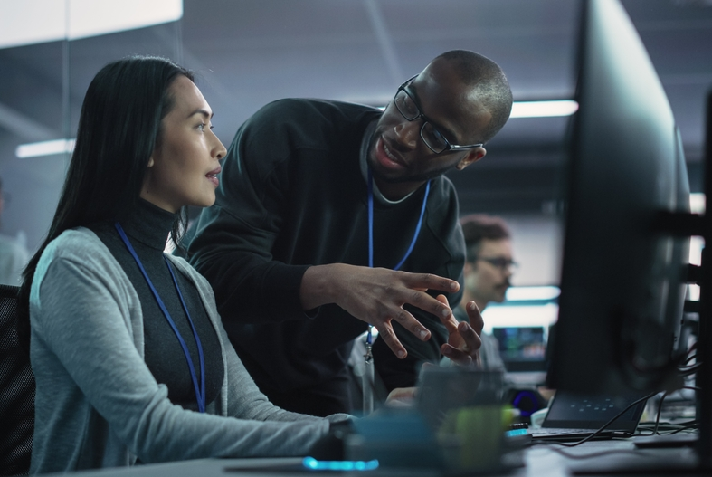 A woman sits at a computer station, engaged in conversation with a man standing next to her.