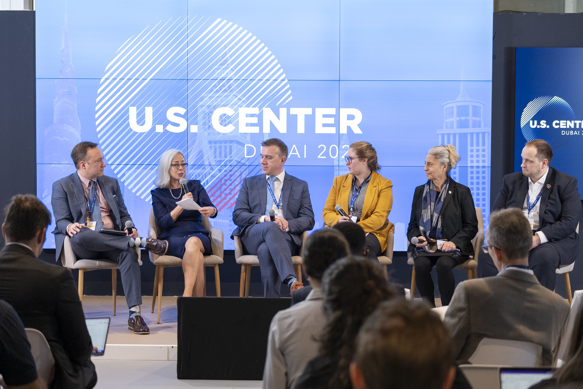 Thea Lee holds a microphone while speaking during a panel discussion with five other people during COP28. The backdrop says “U.S. Center, Dubai 2023.”