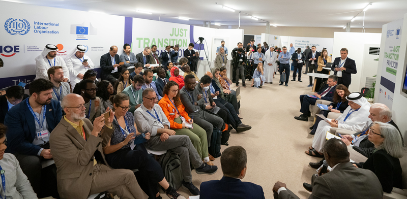 Wide-angle shot of the Just Transition pavilion. Thea Lee sits on a panel with several other people while a small audience looks on and staff film the discussion. Many people are taking notes.