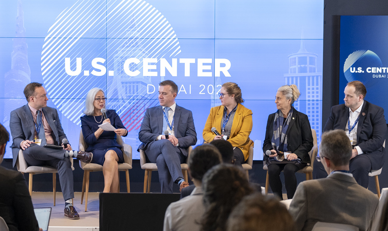 Thea Lee holds a microphone while speaking during a panel discussion with five other people during COP28. The backdrop says “U.S. Center, Dubai 2023.”