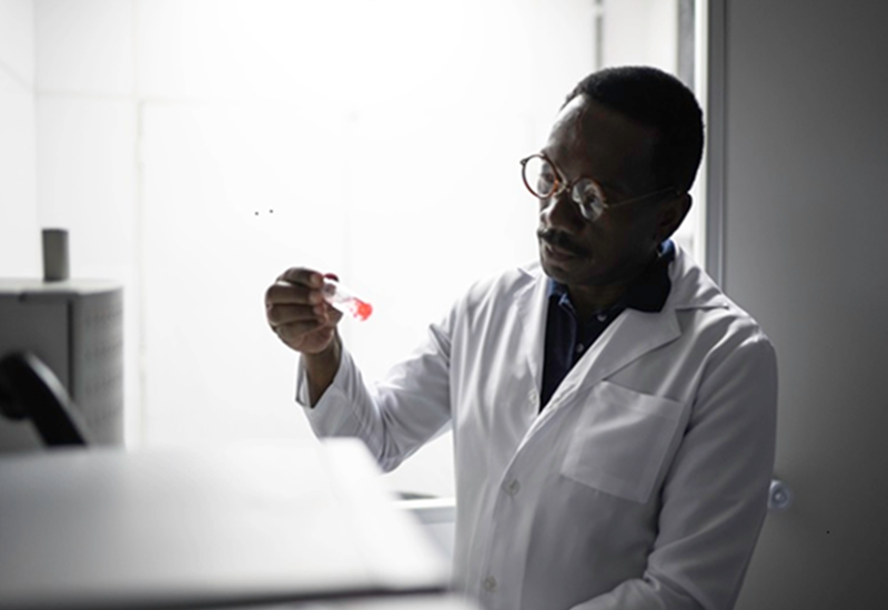 A black scientist in a white lab coat examines a test tube.