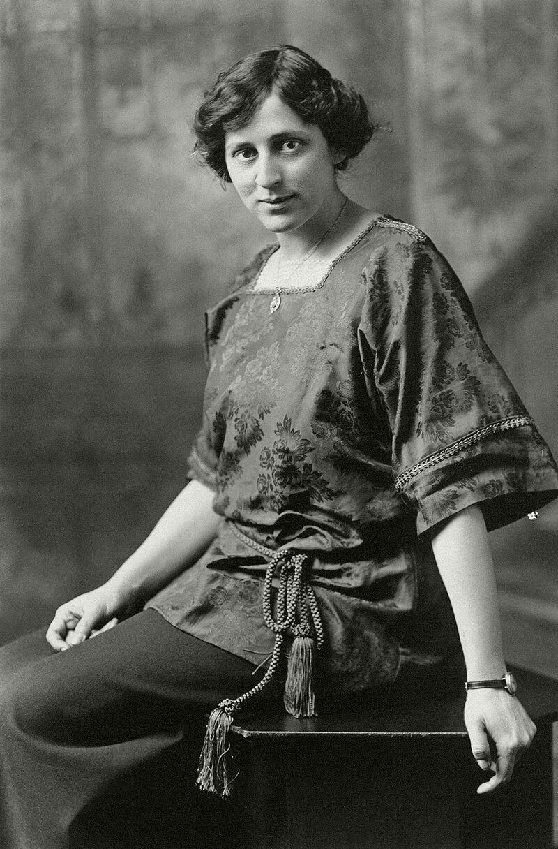 Black and white photo of a woman wearing Victorian-era clothes, sitting on a bench.