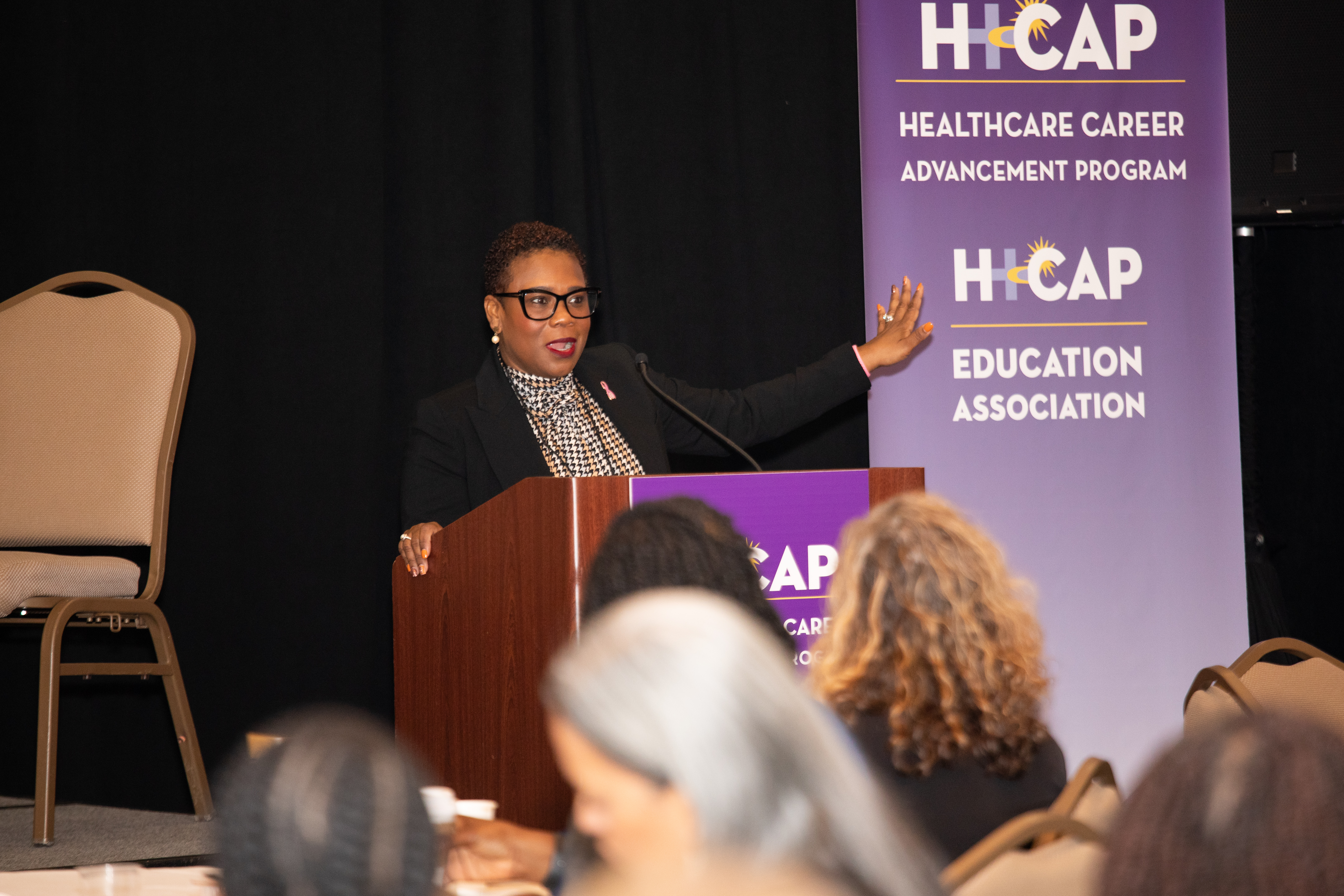 A woman in a dark jacket and glasses speaks at a podium. A banner in the background reads
