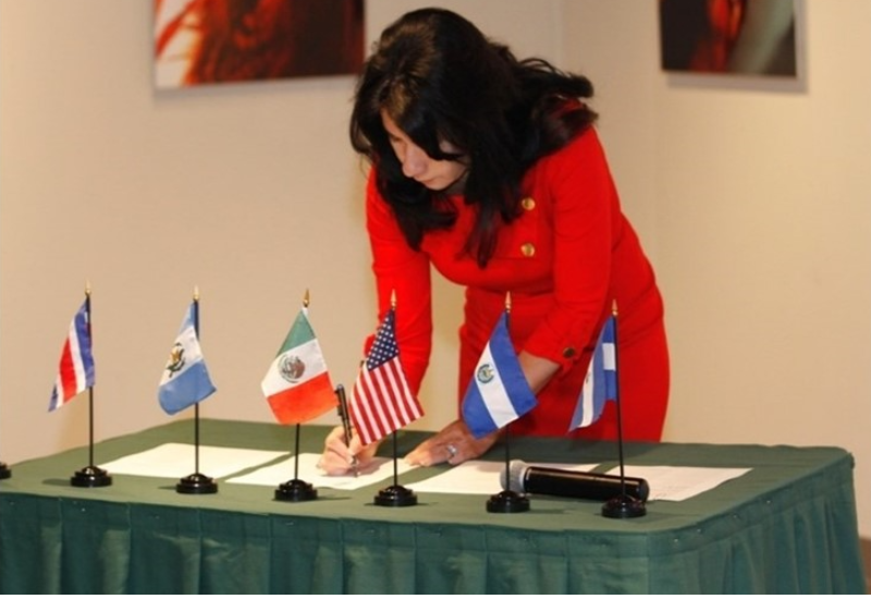 A woman in a red suit signs a document on a table with small flags representing the U.S. and several other countries.