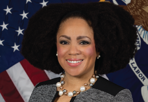 Pictured is a black woman with shoulder length natural hair smiling at the camera in front of the American and U.S. Department of Labor flags. 