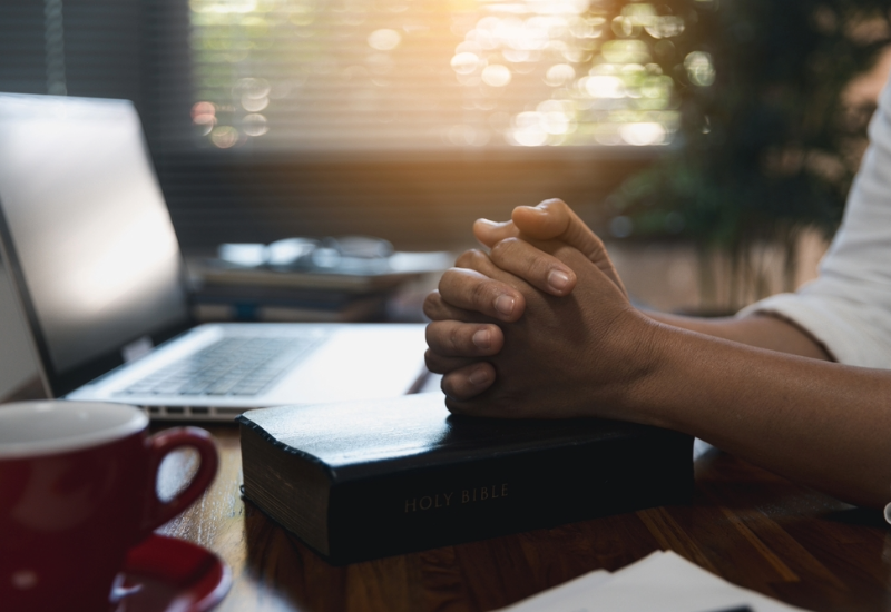 Close-up of a desktop with a laptop, mug and bible. A person sits with their hands clasped over the bible.